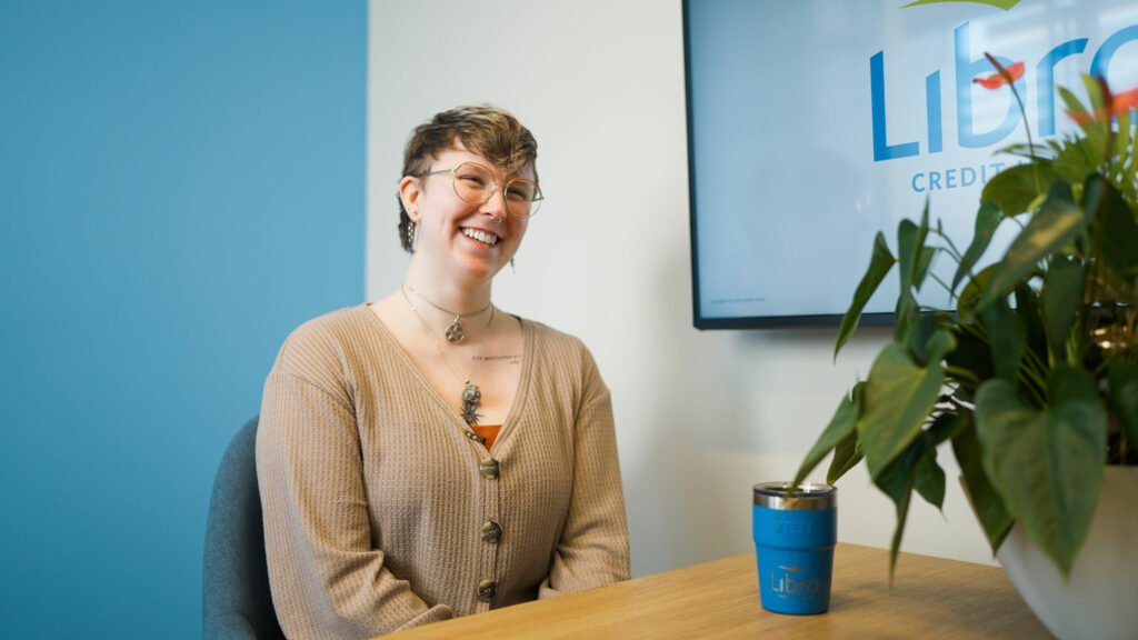 A person with short hair and glasses sits at a desk, smiling, with a mug and a plant in the foreground and a 