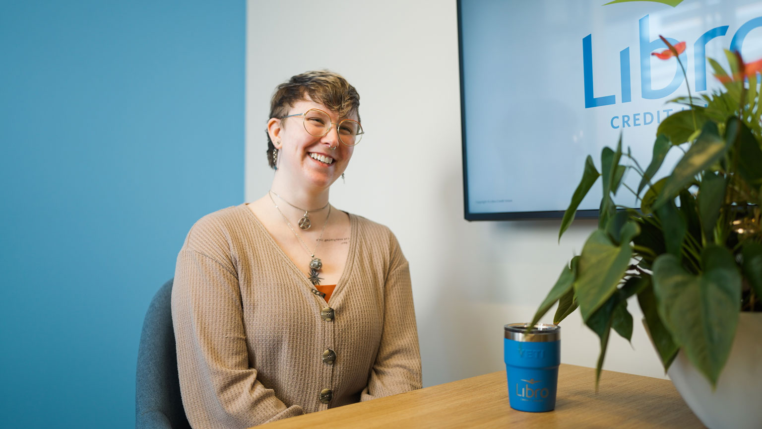 A person with short hair and glasses sits at a desk, smiling, with a mug and a plant in the foreground and a 