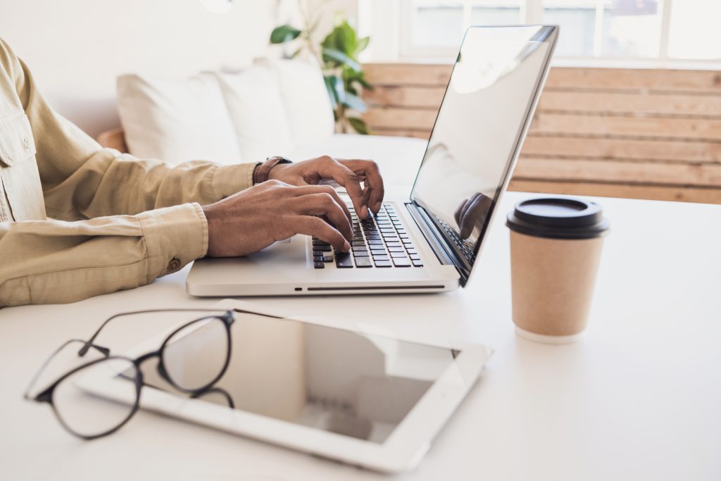 Hands typing on a laptop at a table with glass coffee and paper nearby.