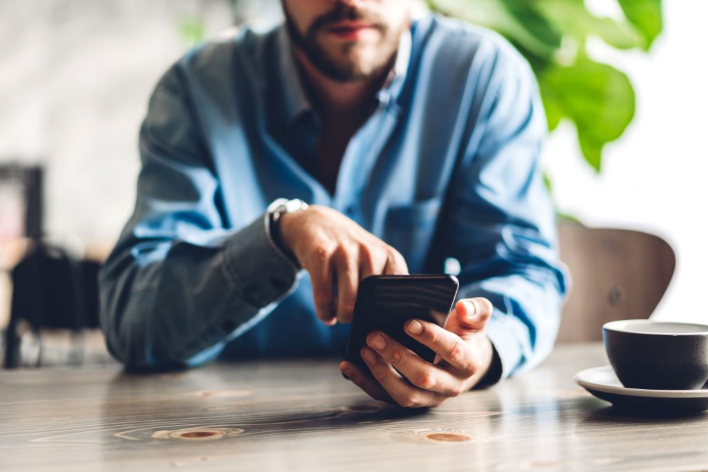 A man in a blue shirt sits at a table, using a smartphone with one hand. A cup and saucer are on the table in front of him.