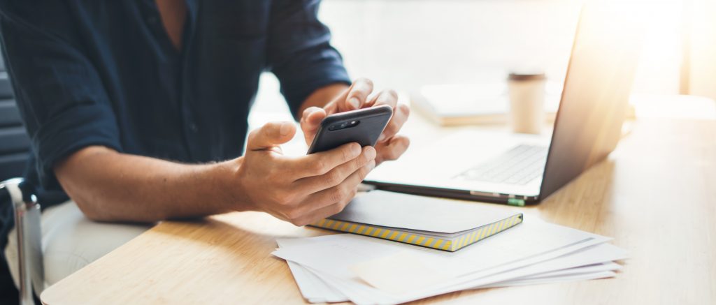 Person using a smartphone at a desk with a laptop, notebook, and papers, suggesting a work or study environment.