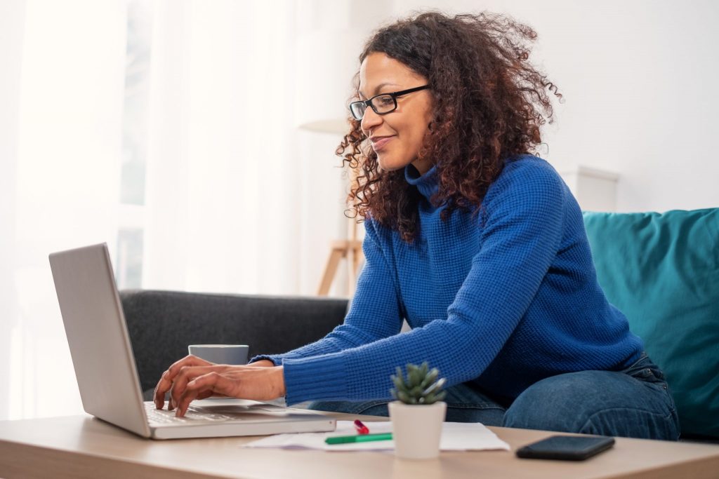 A person wearing a blue sweater sits on a couch and types on a laptop at a coffee table, with documents, a pen, a phone, and a small plant nearby.
