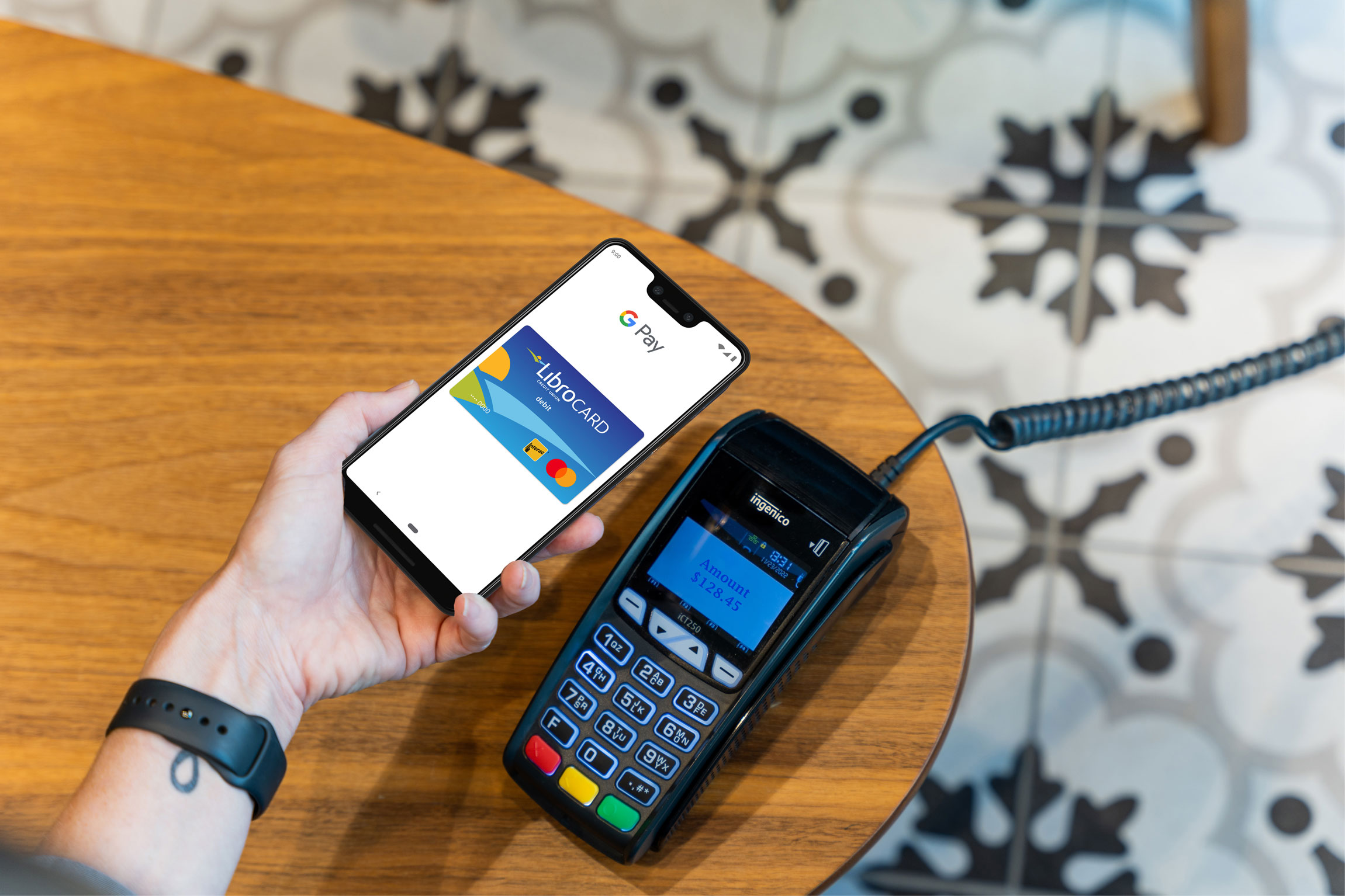 A person holds a smartphone with a Google Pay screen near a card payment terminal on a wooden table, preparing for a contactless transaction.