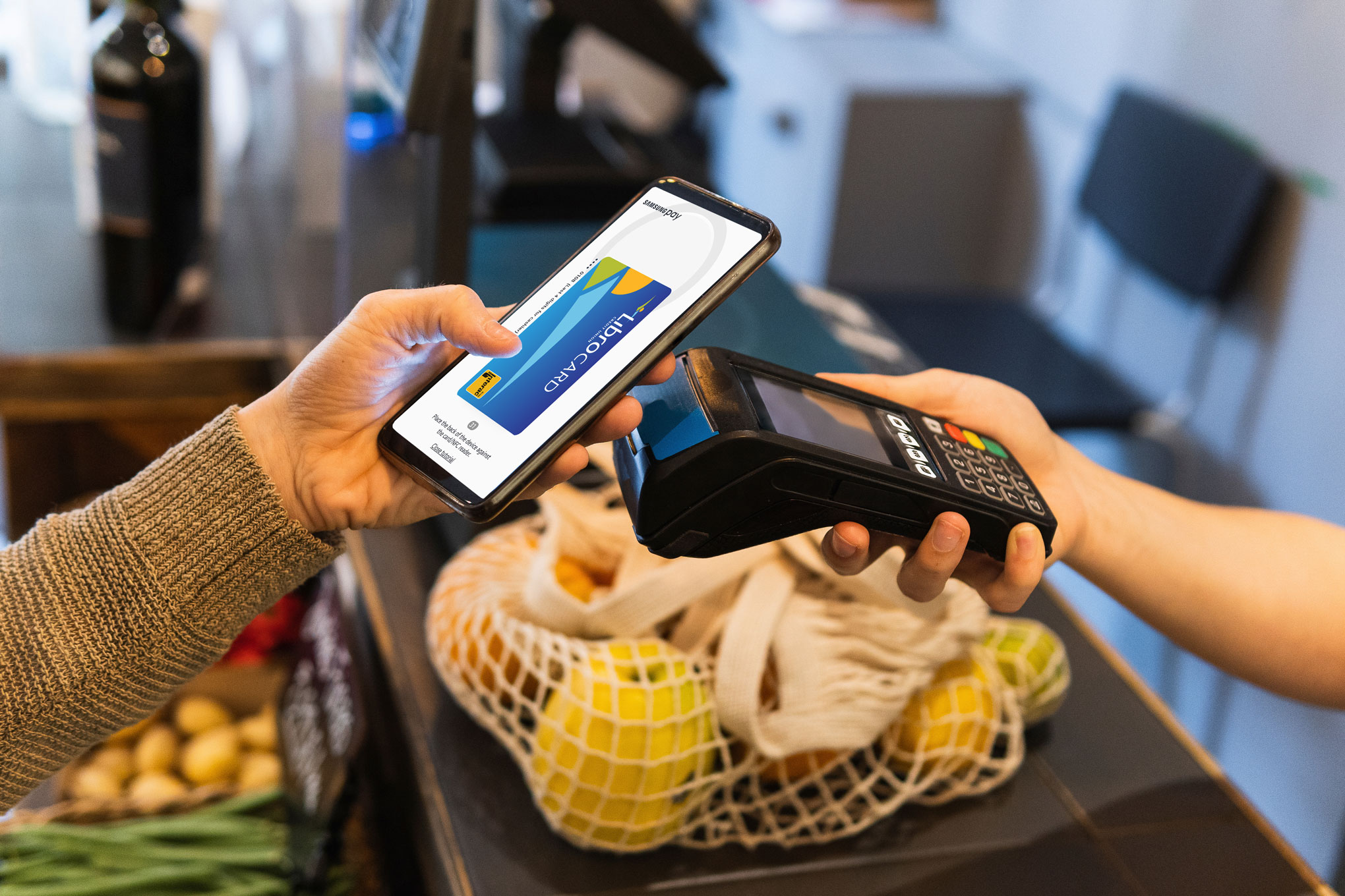 A person uses a smartphone with a digital wallet app to make a contactless payment at a card reader near a reusable shopping bag with groceries.