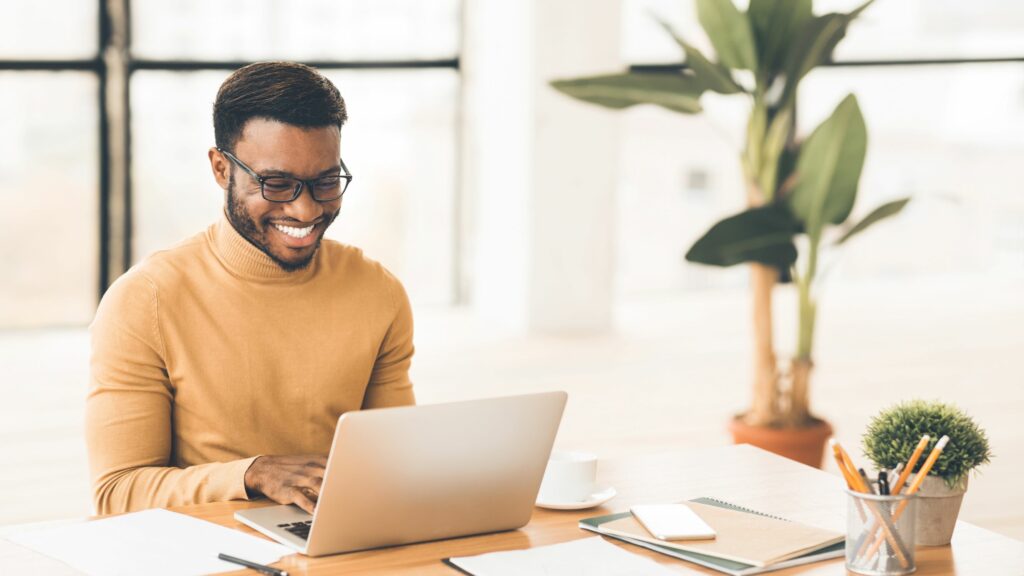 A man wearing glasses and a tan sweater smiles while working on a laptop at a desk with a plant, coffee cup, and office supplies in a bright modern office.