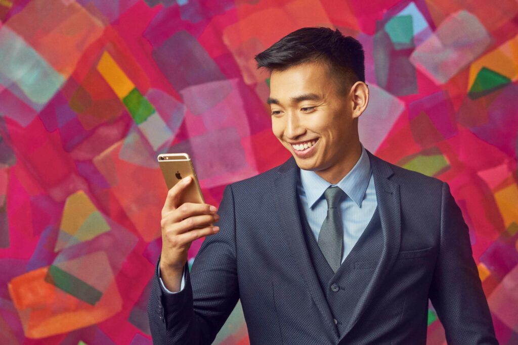 A man in a blue suit smiles while looking at his smartphone, standing in front of a colorful geometric background.