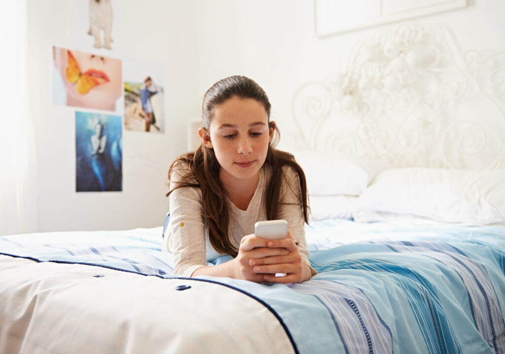 A girl lies on her bed in a bright room, looking at a smartphone. There are posters on the wall and the bed is covered with a blue-striped blanket.