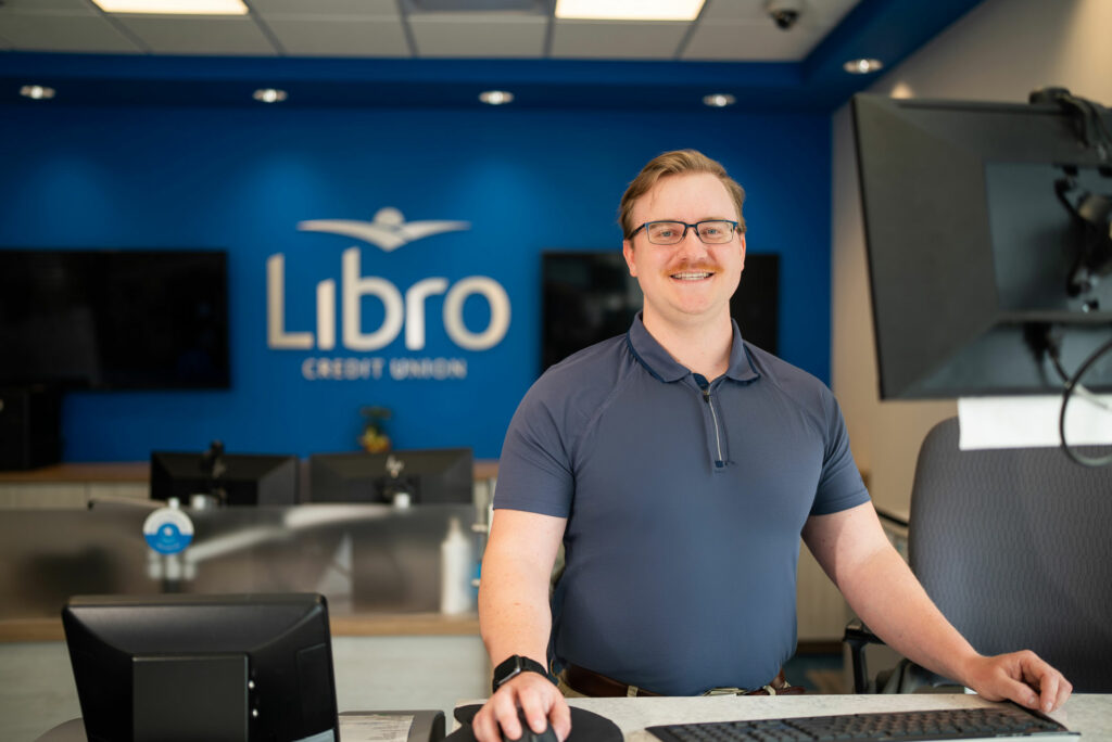 A man in a blue shirt stands smiling at a workstation in a modern office with a 