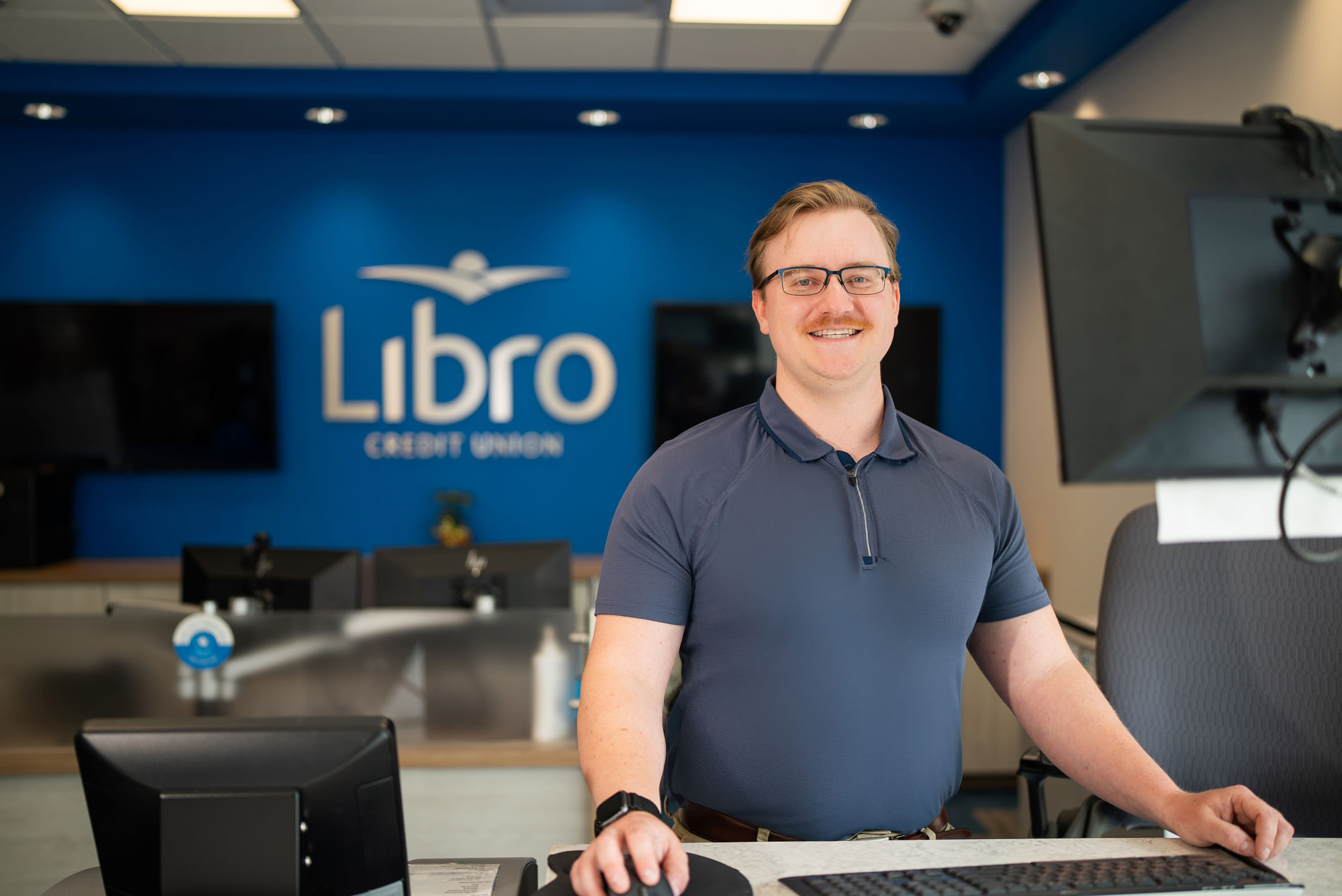 A man in a blue shirt stands smiling at a workstation in a modern office with a 