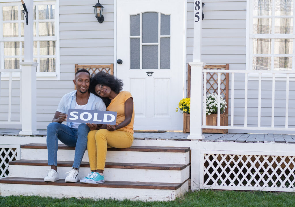 Male and female sitting on porch of house holding a small rectangular sold sign