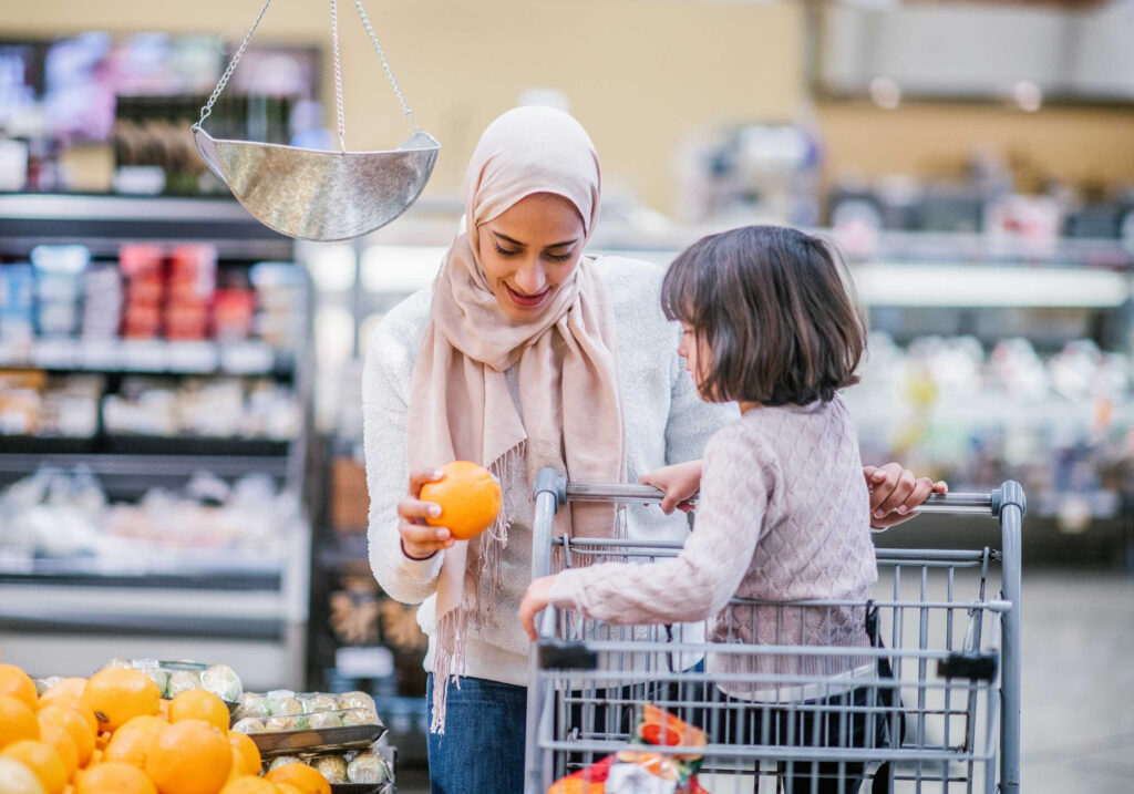 A woman in a hijab selects an orange while shopping with a young girl sitting in a grocery cart in the produce section of a supermarket.