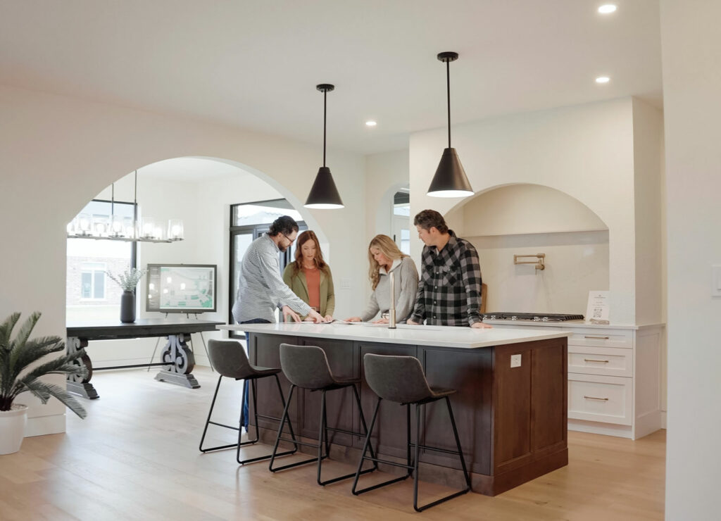 Four people stand around a kitchen island looking at documents in a modern, bright kitchen with pendant lights and barstools.