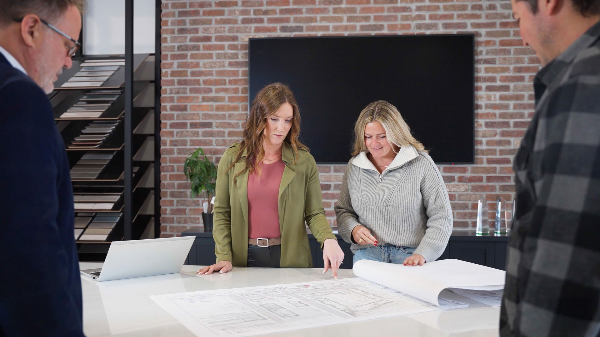 Four people stand around a table reviewing architectural blueprints in a modern office with shelves, a TV, and a laptop visible in the background.