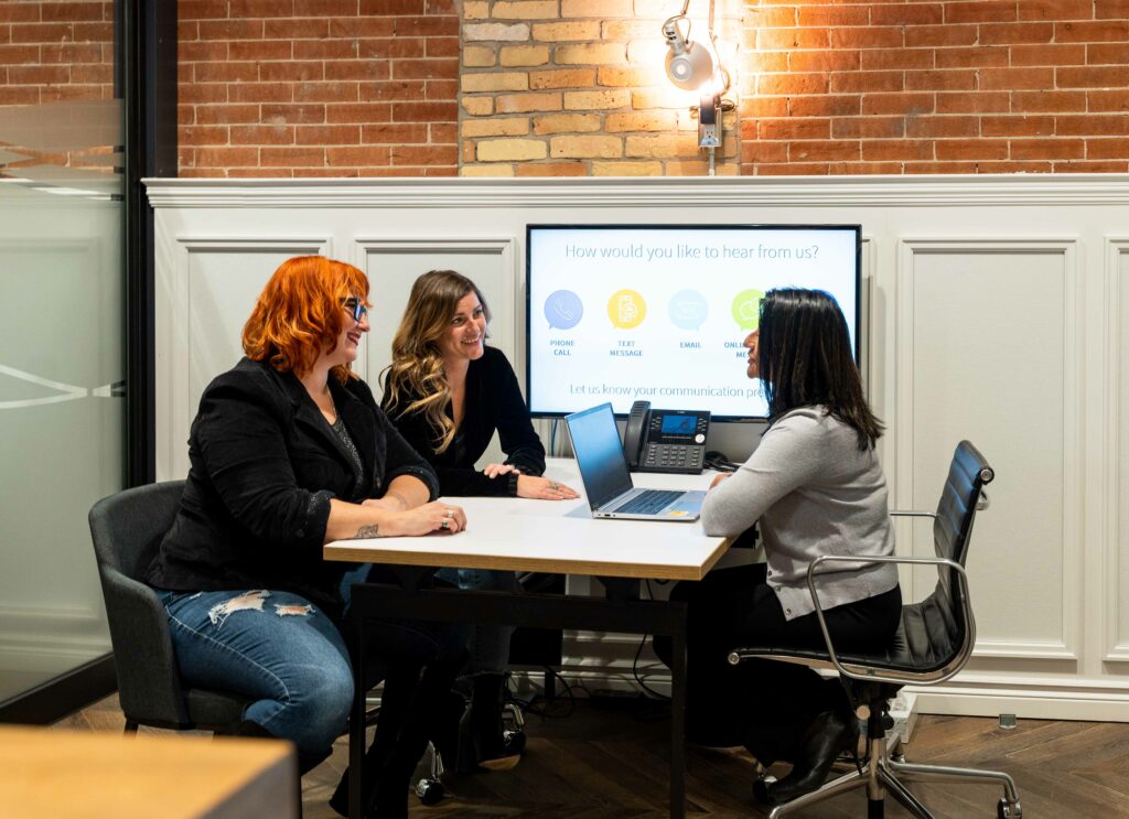 Two females speaking with another female sitting across a desk from each other.