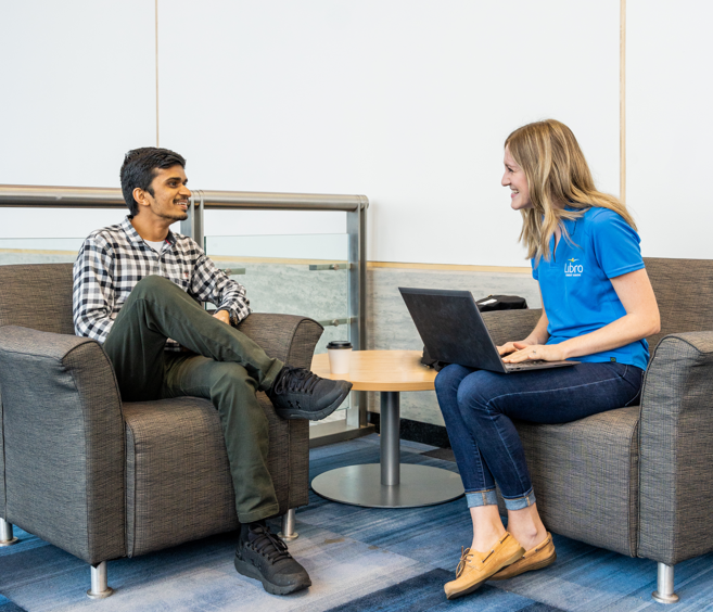 Two people sit in armchairs having a conversation; one holds a laptop and wears a blue shirt, while the other wears a plaid shirt and has legs crossed—discussing Libro’s banking and financial services in Ontario.