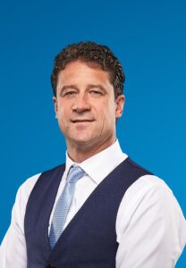 A man with short dark hair wearing a white shirt, blue vest, and light blue tie poses against a plain blue background.