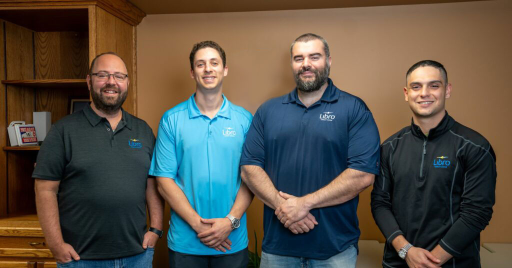 Four men stand side by side indoors, all wearing shirts with the 