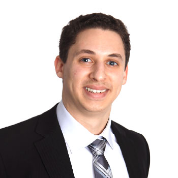 A man in a black suit, white shirt, and patterned tie smiles at the camera against a plain white background.