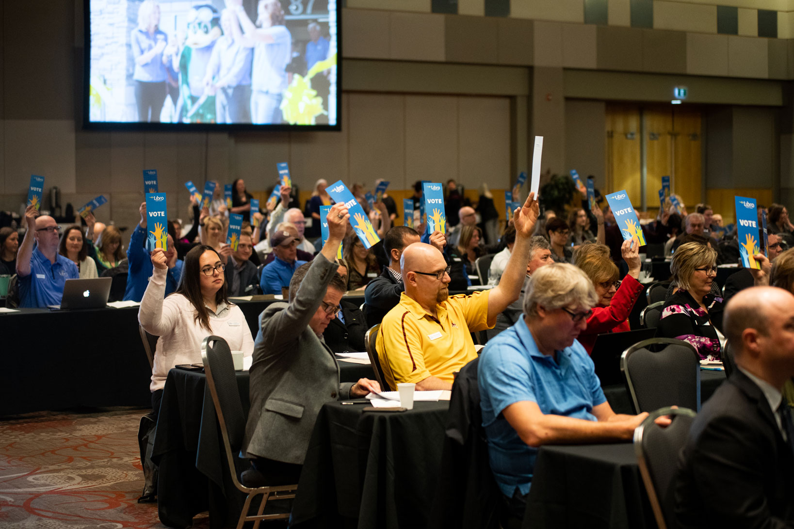 The audience of Libro Owners at an annual general meeting, sitting in chairs and some holding up signs that say 