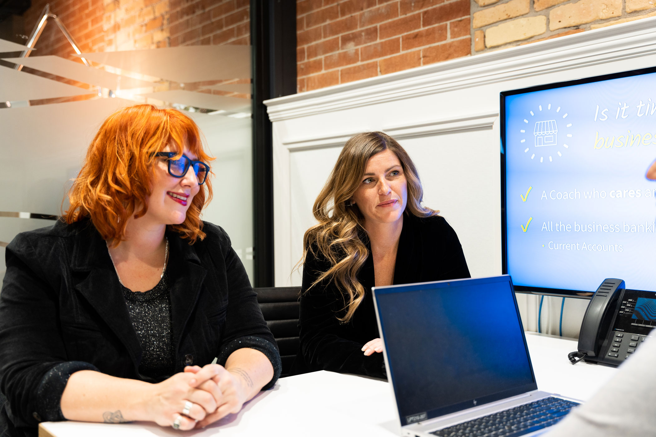 Two women sit at a table in an office, facing a laptop. A presentation slide on a monitor behind them displays business-related information.