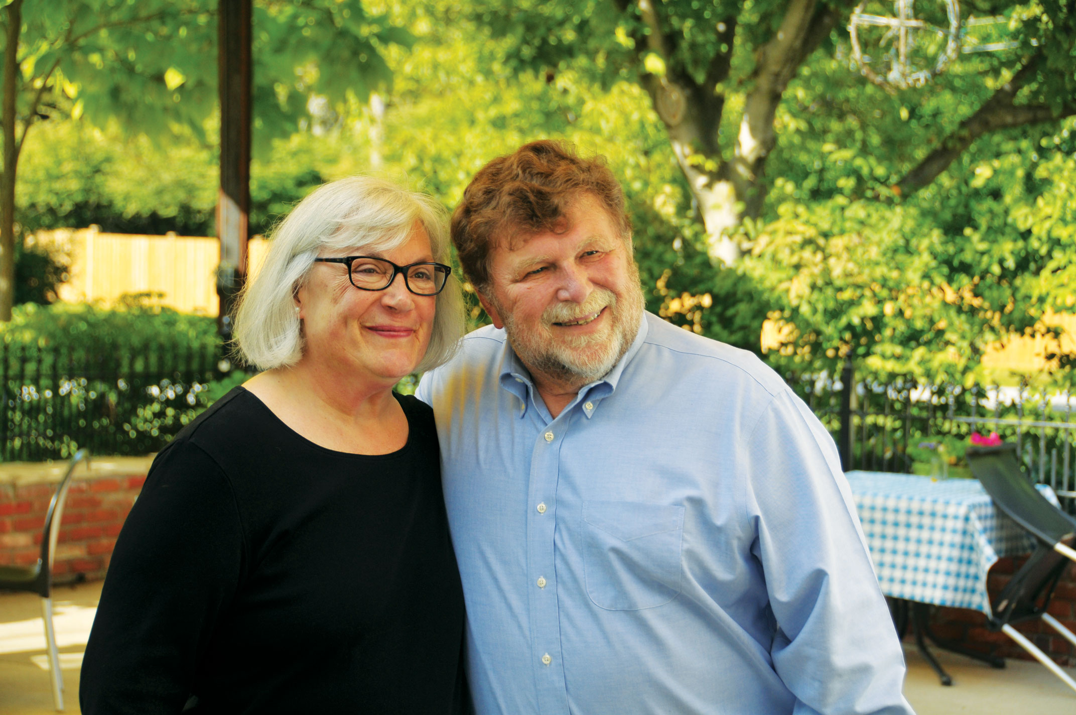 An older woman and man stand close together outdoors, smiling at the camera. Trees and a table with a checkered cloth are visible in the background.