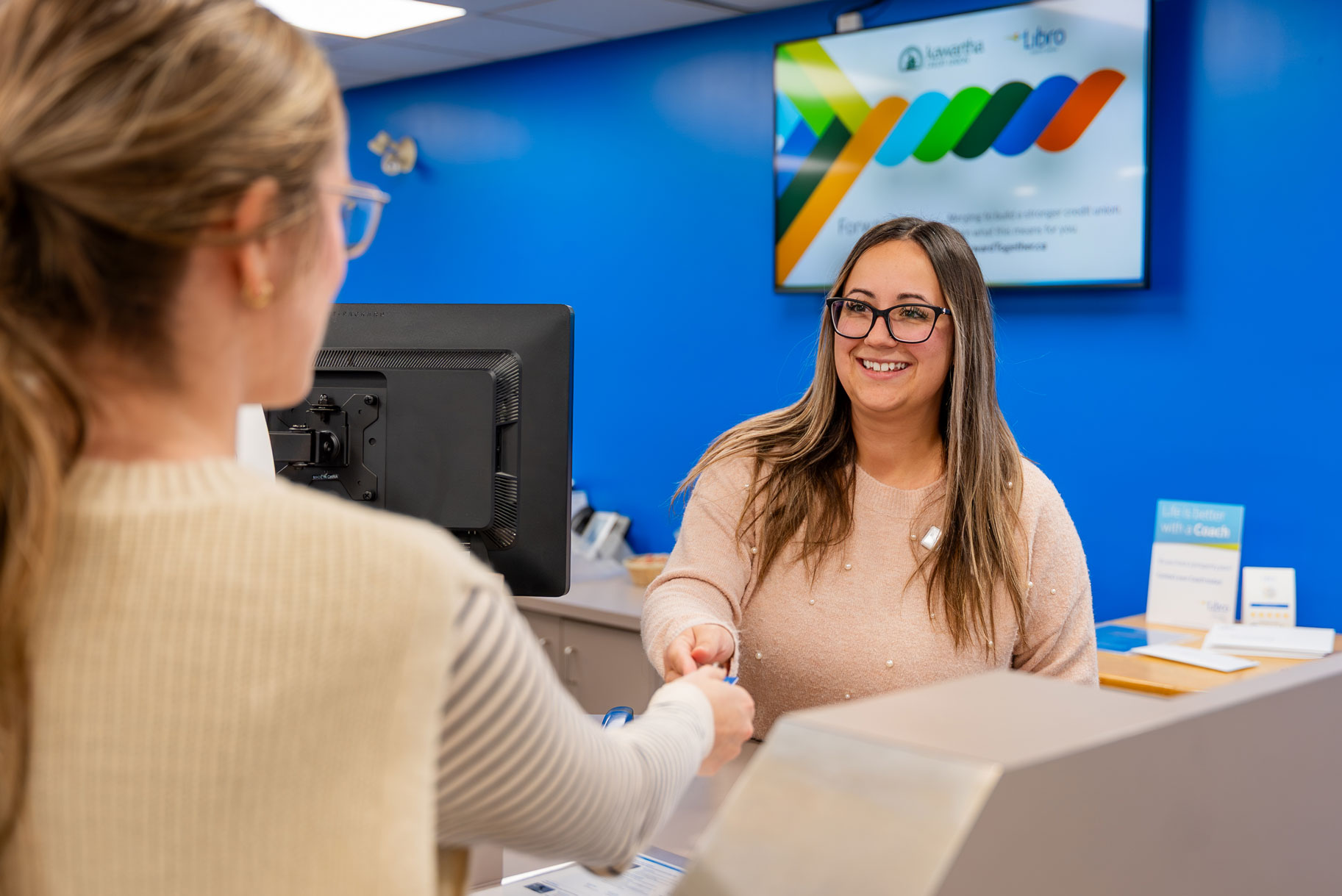 A woman at a service counter hands a card to a smiling receptionist in an office with a blue wall and informational display.