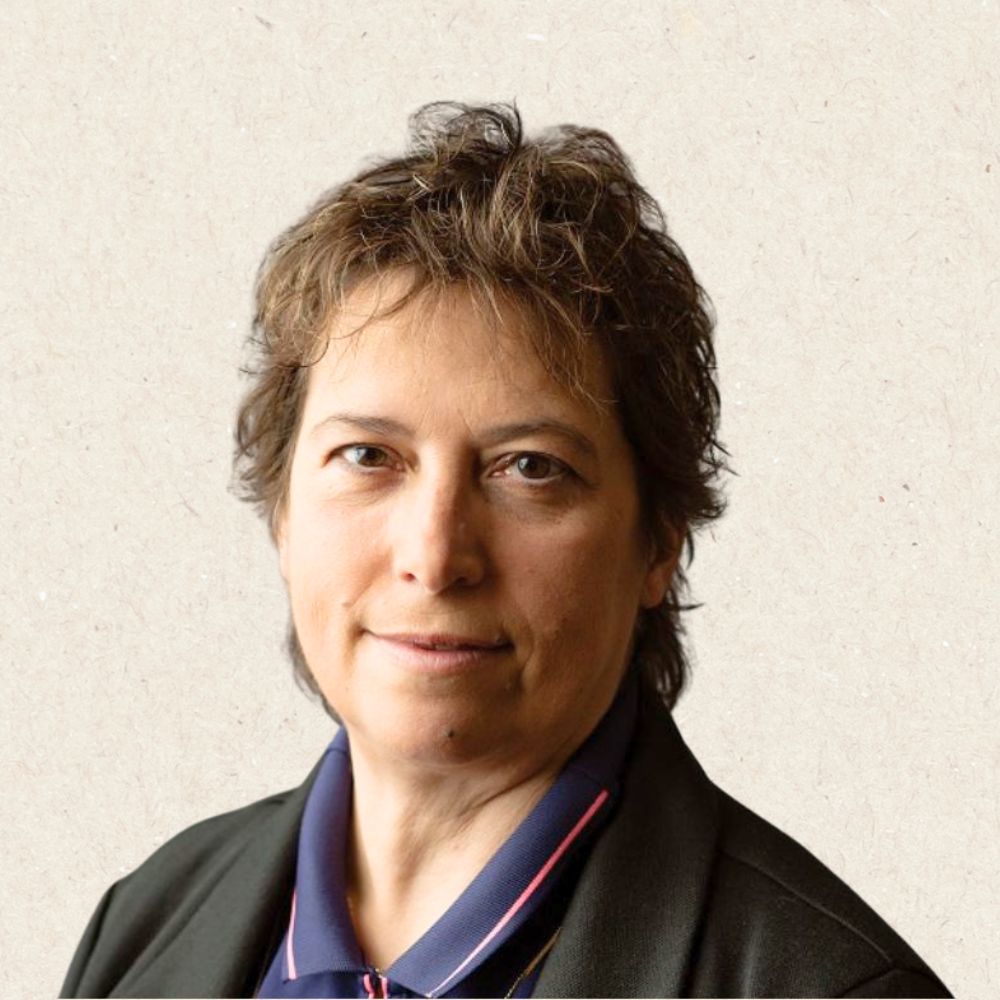 A person with short, wavy brown hair wearing a dark blazer and a collared shirt poses in front of a neutral background.