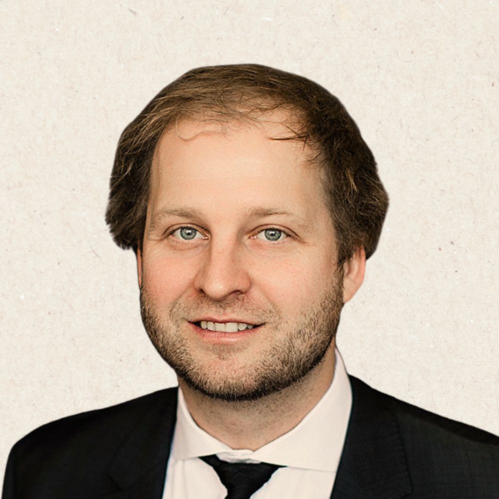 A man with light skin, short brown hair, and blue eyes wearing a black suit, white shirt, and black tie, smiling at the camera against a light background.