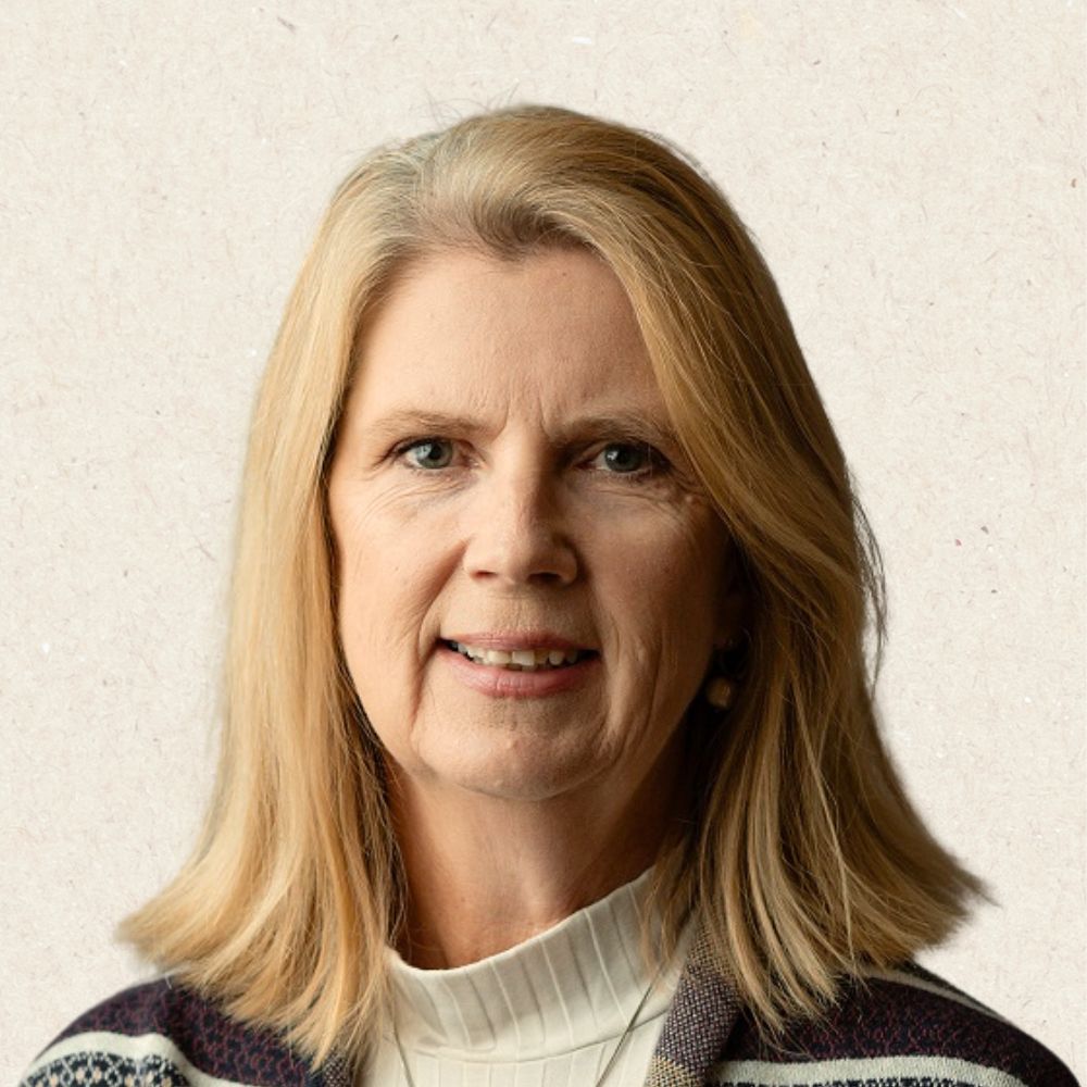 A middle-aged woman with straight blonde hair, wearing a white turtleneck sweater and patterned cardigan, faces the camera against a plain light background.