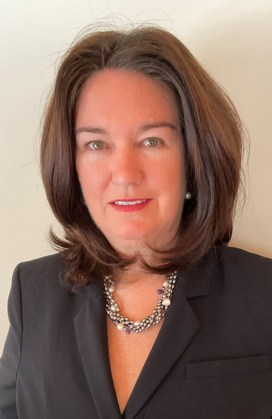 Woman with medium-length brown hair wearing a black blazer and a beaded necklace, standing against a plain light background.
